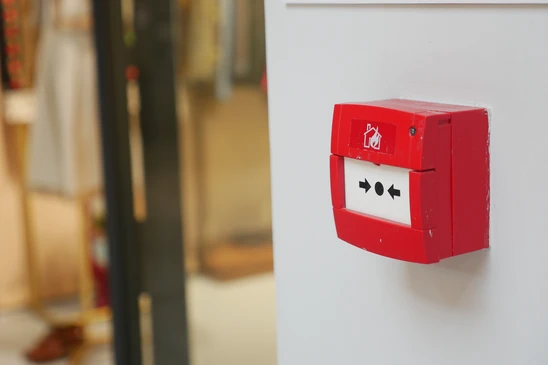 Gros plan sur un déclencheur manuel d'alarme incendie rouge vif fixé au mur blanc, en angle légèrement surplombant. Le boîtier affiche le pictogramme de flamme normalisé blanc et les inscriptions "ALARME INCENDIE" en noir. Le vitre cassable transparente protégeant le bouton d'actionnement est visible. En arrière-plan très flou, un couloir professionnel lumineux rappelant un environnement commercial ou tertiaire. Éclairage naturel, photographie nette en détail sur l'équipement.